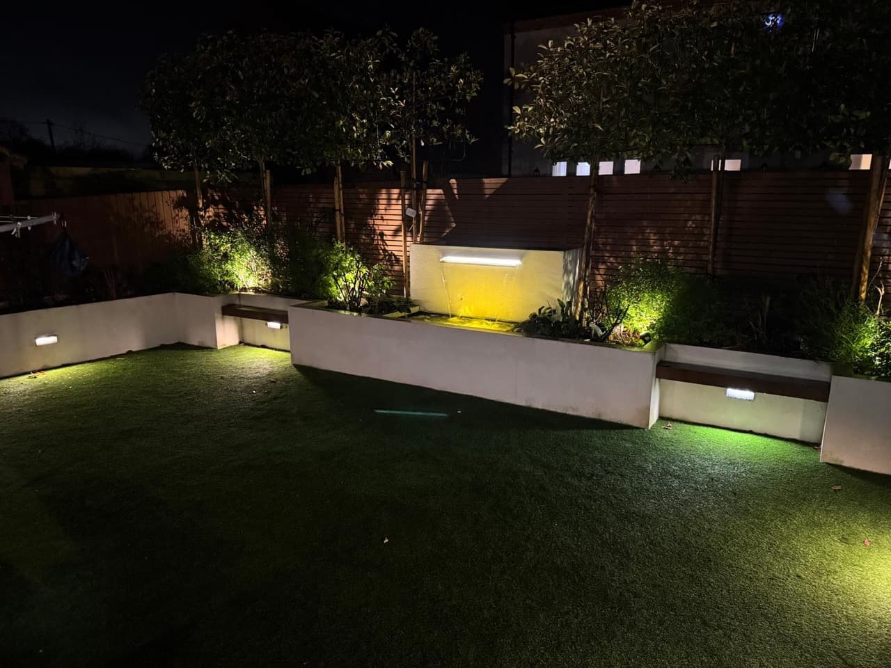 Garden at night with a lit water feature, uplit raised beds and recessed wall lights along the lawn